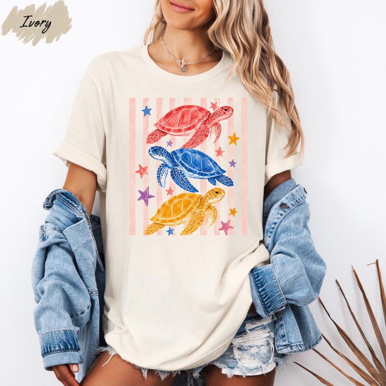 A woman wearing a white t-shirt with a colorful graphic of three sea turtles and stars, paired with denim shorts and a denim jacket.