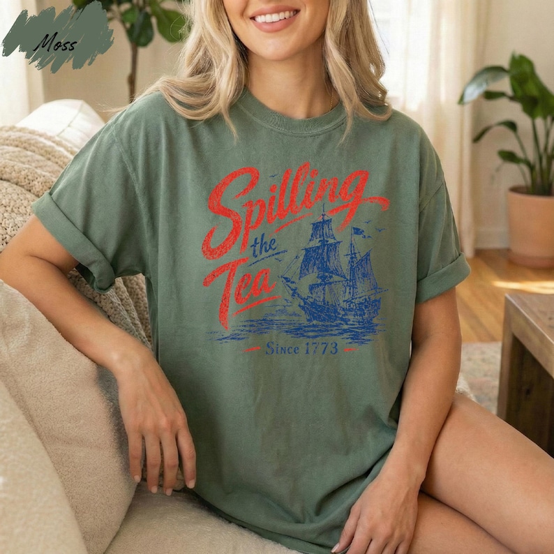 A woman wearing a green t-shirt with a graphic design of a ship and the text "Spilling the Tea Since 1773" is sitting on a couch.