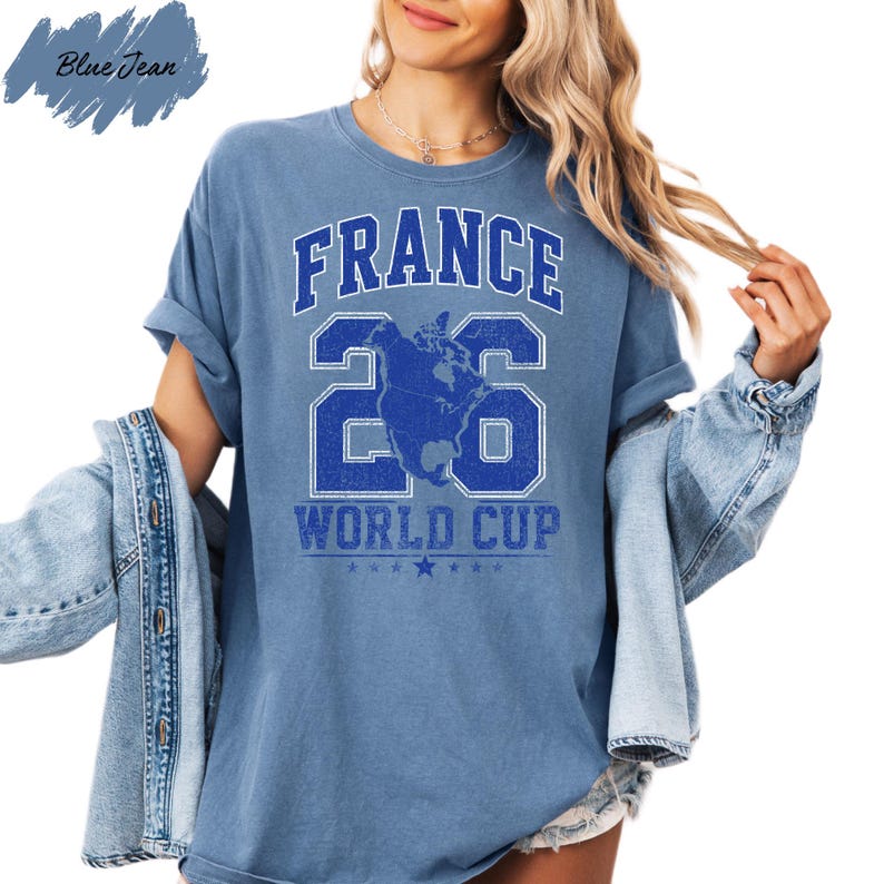 A woman wearing a blue t-shirt with the text "FRANCE 26 WORLD CUP" printed on it, along with a graphic of a globe. She is also wearing a denim jacket and has her hair styled in loose waves.