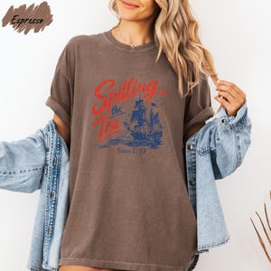 A woman wearing a brown t-shirt with a graphic design of a ship and the text "Sailing the Tea" printed on it, along with the text "Since 1773". She is also wearing a denim jacket and has her arms outstretched.