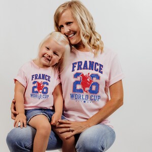 A smiling woman wearing a pink t-shirt with the text "FRANCE 26 WORLD CUP" and a young girl wearing a matching t-shirt.