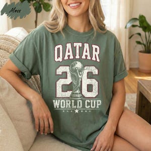 A woman wearing a green t-shirt with the text "QATAR 26 WORLD CUP" printed on it, sitting on a couch.
