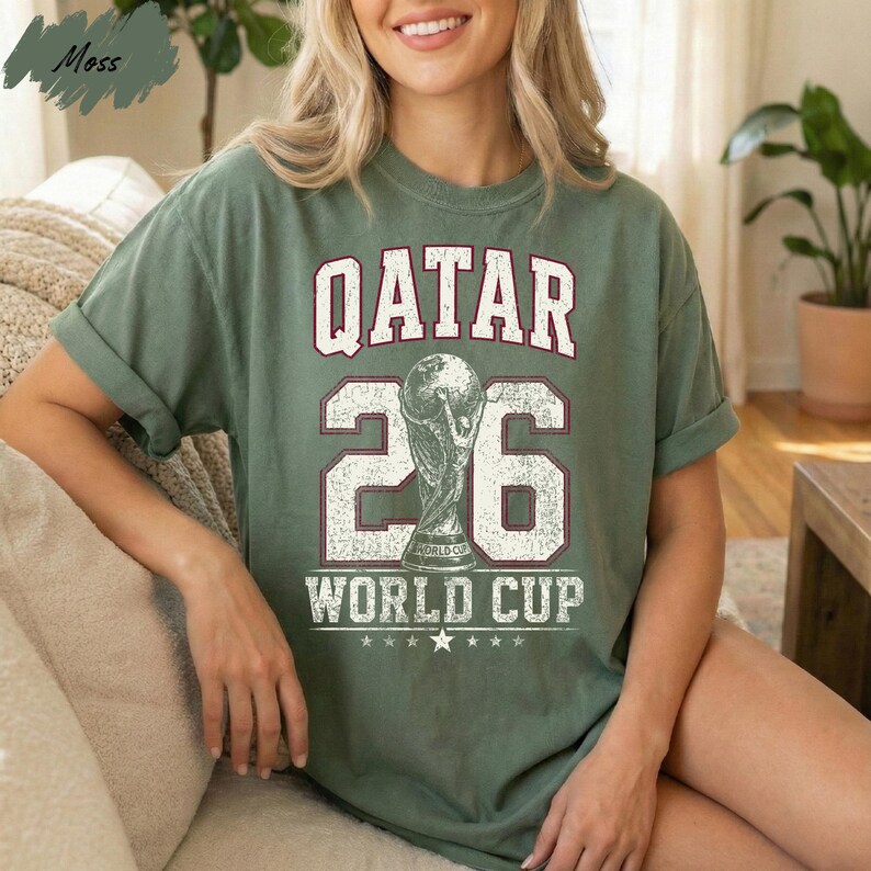 A woman wearing a green t-shirt with the text "QATAR 26 WORLD CUP" printed on it, sitting on a couch.