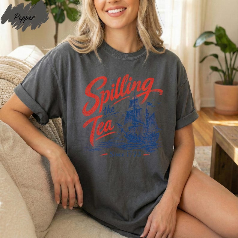 A woman wearing a gray t-shirt with a graphic design of a ship and the text "Spilling the Tea Since 1730" is sitting on a couch.