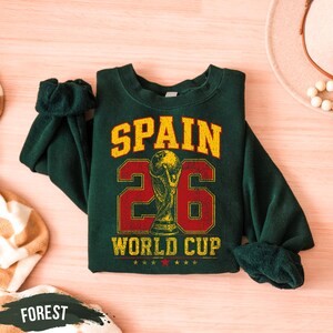 A green sweatshirt with the text "SPAIN 26 WORLD CUP" printed on it, along with a trophy icon, is displayed on a wooden surface.
