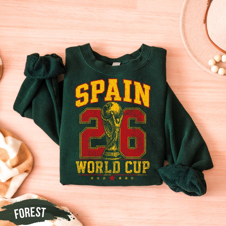 A green sweatshirt with the text "SPAIN 26 WORLD CUP" printed on it, along with a trophy icon, is displayed on a wooden surface.