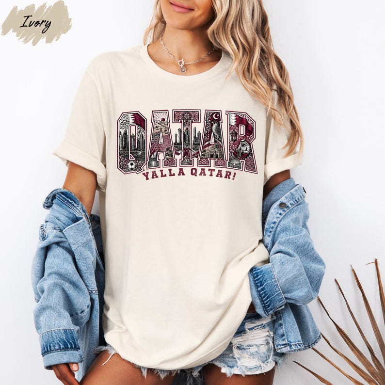 A woman wearing a white t-shirt with the word "QATAR" printed on it, along with the phrase "YALLA QATAR!" printed below. She is also wearing denim clothing, including a denim jacket and denim shorts.