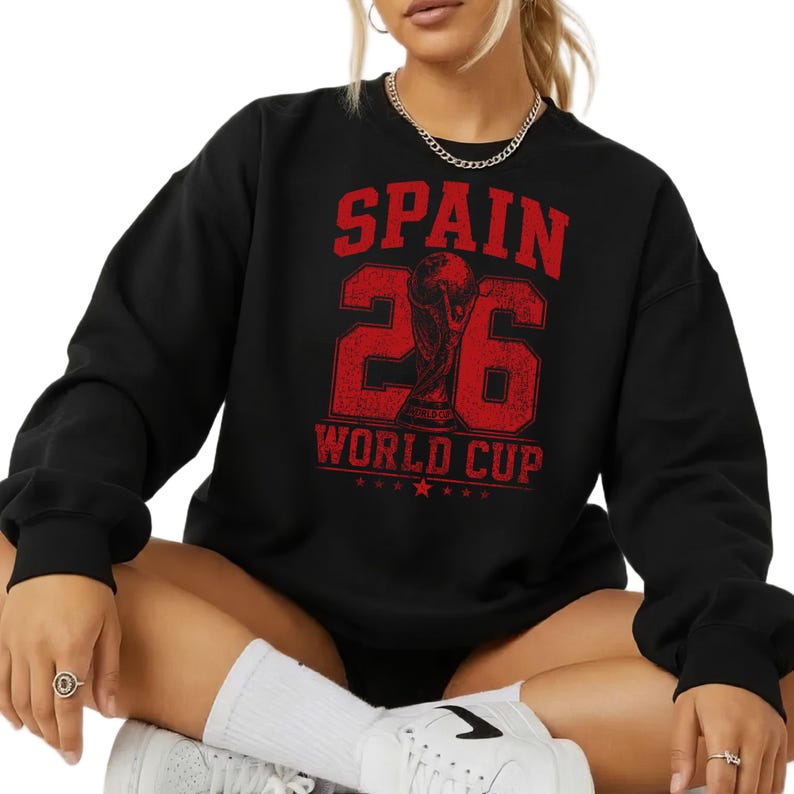 A person wearing a black sweatshirt with a red and white graphic design that says "SPAIN 26 WORLD CUP" printed on it.