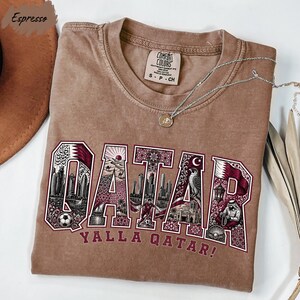 a brown t-shirt with the word "QATAR" printed on it, along with various illustrations of landmarks and symbols associated with Qatar.