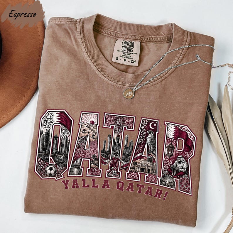 a brown t-shirt with the word "QATAR" printed on it, along with various illustrations of landmarks and symbols associated with Qatar.