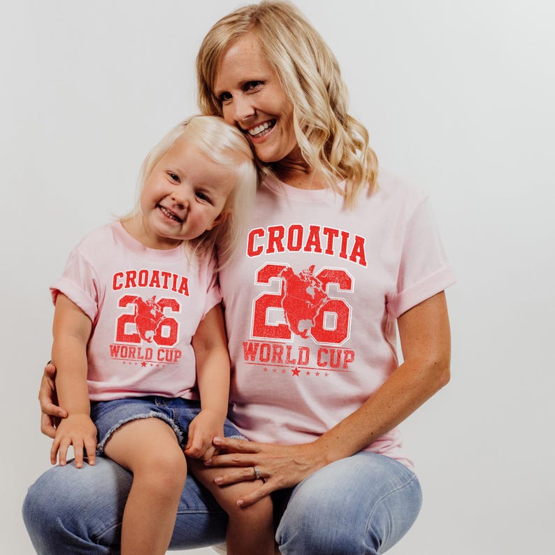 A smiling woman wearing a pink t-shirt with the word "Croatia" and the number "26" is holding a young girl in a matching t-shirt.