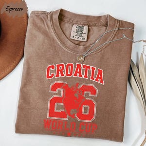 A brown t-shirt with the text "Croatia" and "World Cup" printed on it, along with the number "26". The t-shirt is laid out on a white surface, with a brown hat and some feathers visible in the background.