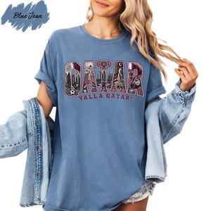 A woman wearing a blue t-shirt with the word "QATAR" printed on it, along with the phrase "Yalla Qatar!" printed below. She is also wearing a denim jacket and has her hair styled in loose waves.
