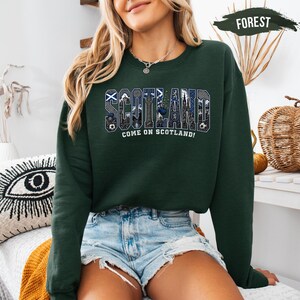A woman wearing a green sweatshirt with the word "SCOTLAND" printed on it, along with the text "COME ON SCOTLAND!" printed below. She is also wearing denim shorts and a necklace. The background includes a wicker basket and a decorative item with an eye design.
