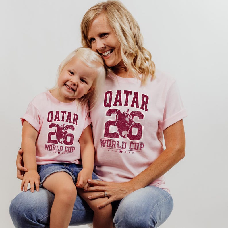 A smiling woman and a young girl wearing matching pink t-shirts with the text "QATAR 26 WORLD CUP" printed on them.
