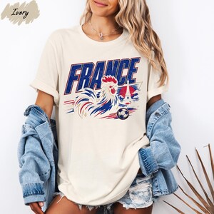 A woman wearing a white t-shirt with a graphic of a rooster and the word "FRANCE" printed on it, along with a denim jacket and denim shorts.