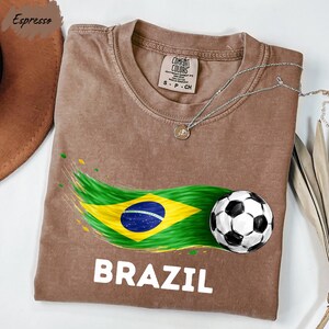 a brown t-shirt with a graphic design depicting the Brazilian flag and a soccer ball, along with the word "BRAZIL" printed on it.