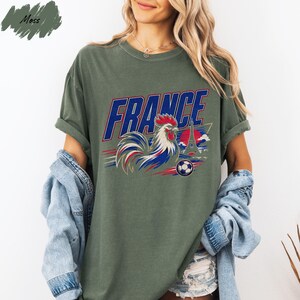 A woman wearing a green t-shirt with a graphic design of a rooster and a soccer ball, along with the word "FRANCE" printed on it. She is also wearing a denim jacket and shorts.