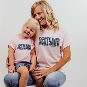 A smiling woman wearing a pink t-shirt with the word "SCOTLAND" and a child wearing a matching pink t-shirt are posing together.