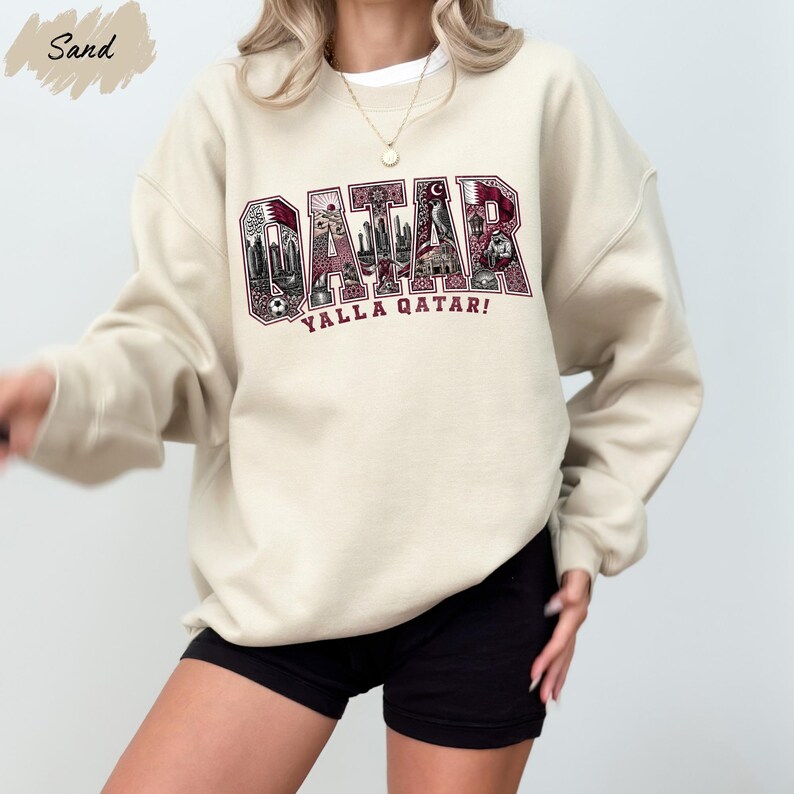 A person wearing a beige sweatshirt with the word "QATAR" printed on it, along with the phrase "YALLA QATAR!" printed below.