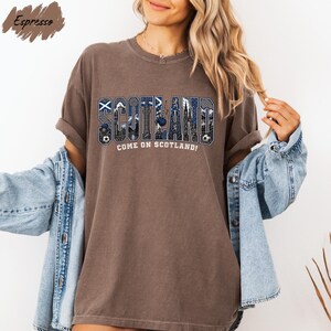 A woman wearing a brown t-shirt with the word "SCOTLAND" printed on it, along with the text "COME ON SCOTLAND!" She is also wearing a denim jacket and has her arms outstretched.