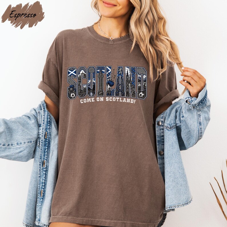 A woman wearing a brown t-shirt with the word "SCOTLAND" printed on it, along with the text "COME ON SCOTLAND!" She is also wearing a denim jacket and has her arms outstretched.