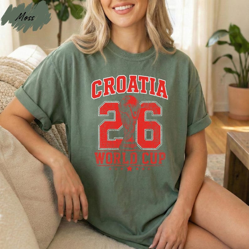 A woman wearing a green t-shirt with the text "Croatia 26 World Cup" printed on it, sitting on a couch.