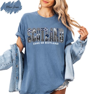 A woman wearing a blue t-shirt with the word "SCOTLAND" printed on it, along with the text "COME ON SCOTLAND!" printed below. She is also wearing a denim jacket and has her hair styled in loose waves.