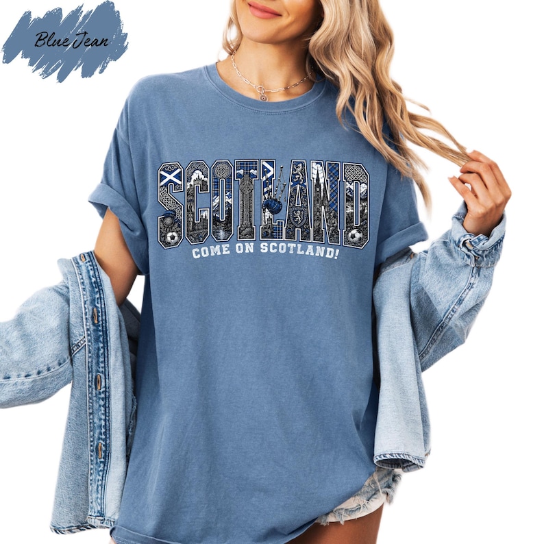 A woman wearing a blue t-shirt with the word "SCOTLAND" printed on it, along with the text "COME ON SCOTLAND!" printed below. She is also wearing a denim jacket and has her hair styled in loose waves.