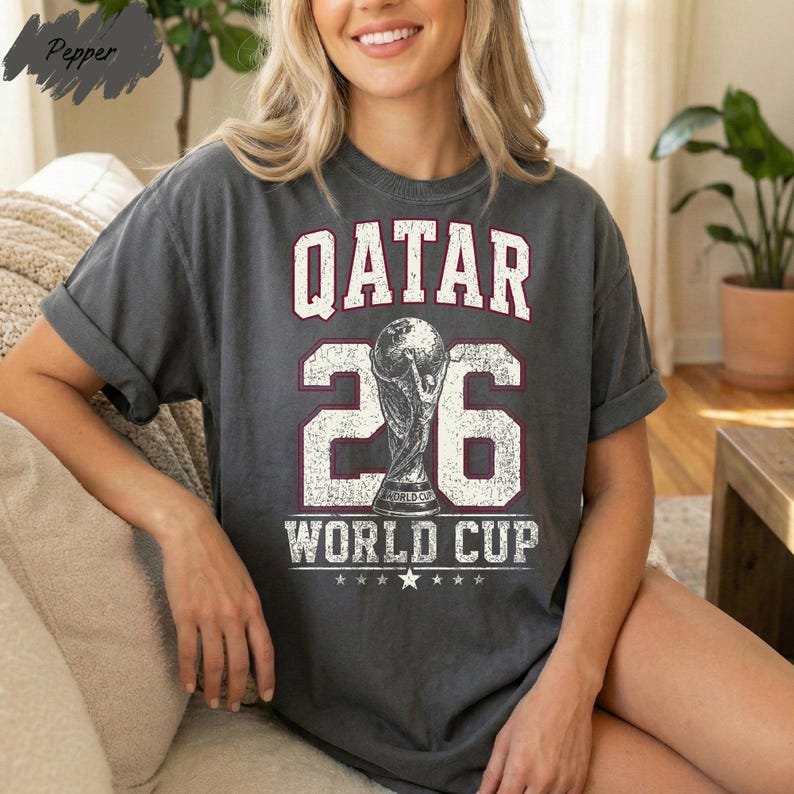 A woman wearing a grey t-shirt with the text "QATAR 26 WORLD CUP" printed on it, sitting on a couch.