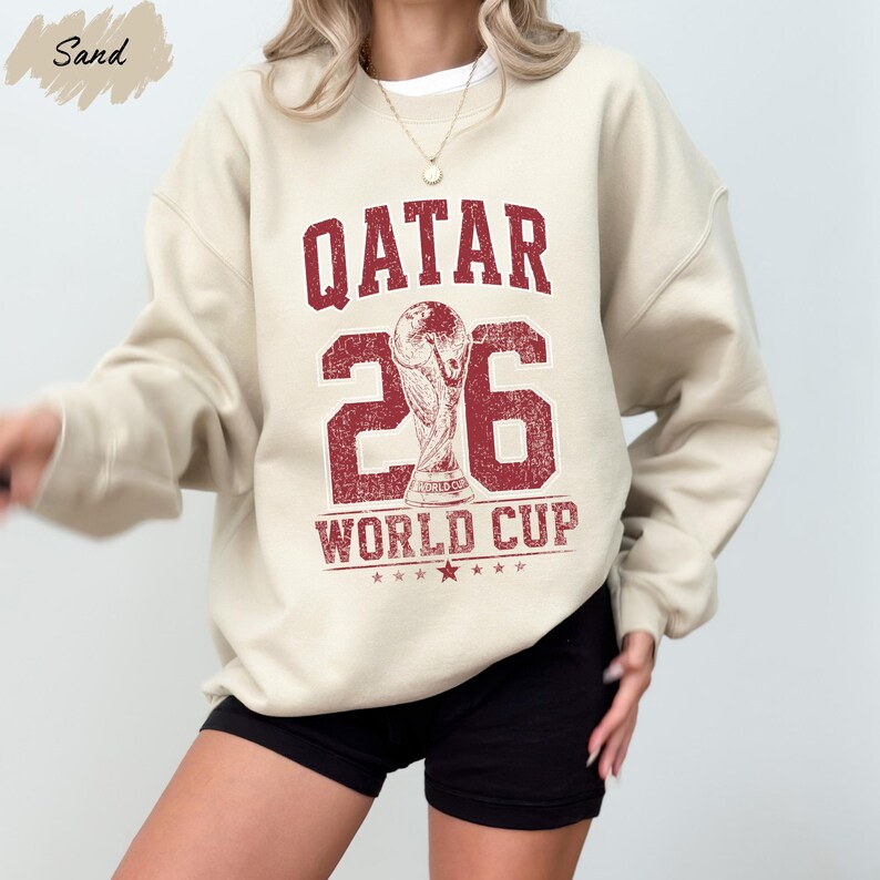 A person wearing a beige sweatshirt with a red and white design that says "QATAR 2006 WORLD CUP".