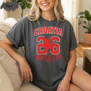 A smiling woman wearing a gray t-shirt with the text "Croatia 26 World Cup" printed on it.