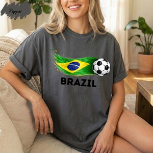 A woman wearing a gray t-shirt with a graphic of the Brazilian flag and a soccer ball, smiling and sitting on a couch.