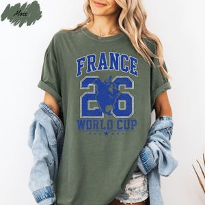 A woman wearing a green t-shirt with the text "FRANCE 26 WORLD CUP" printed on it, standing against a white background.