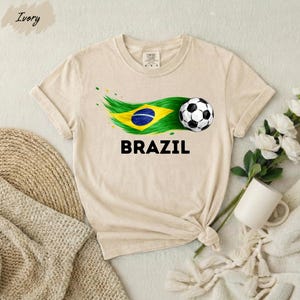 A beige t-shirt with a graphic of a soccer ball and the word "BRAZIL" printed on it, along with a Brazilian flag design. The t-shirt is placed on a white surface, and there are some flowers and a mug nearby.