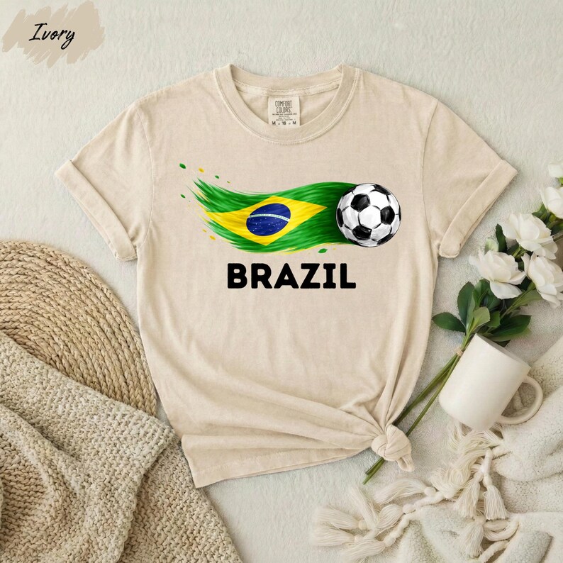 A beige t-shirt with a graphic of a soccer ball and the word "BRAZIL" printed on it, along with a Brazilian flag design. The t-shirt is placed on a white surface, and there are some flowers and a mug nearby.