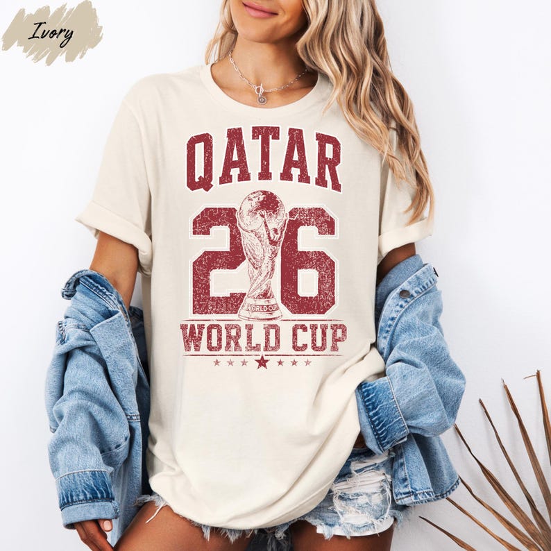 A woman wearing a white t-shirt with the text "Qatar 26 WORLD CUP" printed on it, along with a denim jacket and denim shorts.