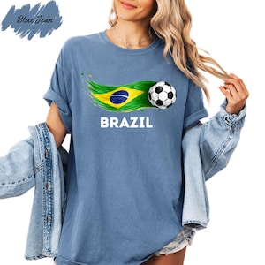 A woman wearing a blue t-shirt with the word "BRAZIL" printed on it, along with a graphic of a soccer ball and the Brazilian flag. She is also wearing a denim jacket and has her hair in a ponytail.