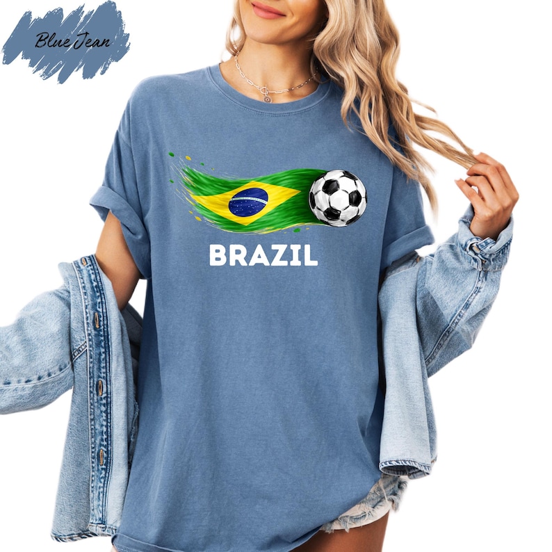 A woman wearing a blue t-shirt with the word "BRAZIL" printed on it, along with a graphic of a soccer ball and the Brazilian flag. She is also wearing a denim jacket and has her hair in a ponytail.