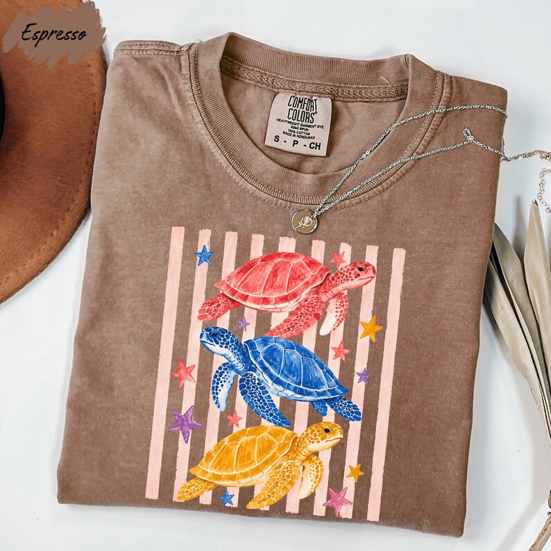 a brown t-shirt with a colorful design of three turtles and stars on it, along with a necklace and a hat.