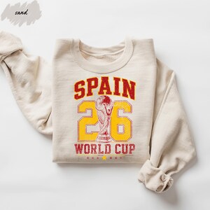A beige sweatshirt with a graphic design featuring the text "SPAIN 26 WORLD CUP" and a trophy icon.