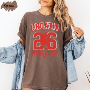A woman wearing a brown t-shirt with the text "CROATIA 26 WORLD CUP" printed on it, along with a graphic of a soccer ball. She is also wearing a denim jacket and has her arms outstretched.
