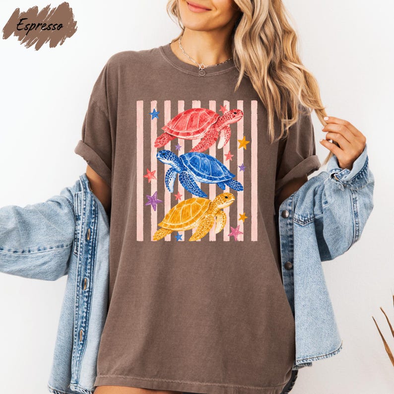A woman wearing a brown t-shirt with a colorful graphic of three sea turtles and stars, along with a denim jacket.