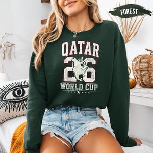 A woman wearing a green sweatshirt with the text "QATAR 26 WORLD CUP" printed on it, paired with denim shorts.