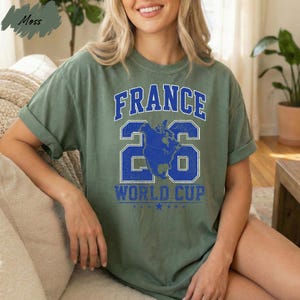 A woman wearing a green t-shirt with the text "FRANCE 26 WORLD CUP" printed on it, sitting on a couch.
