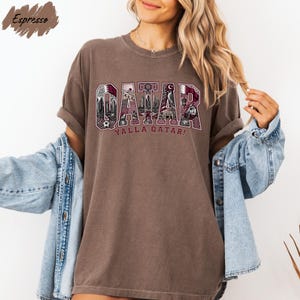 A woman wearing a brown t-shirt with the word "OAKAR" printed on it, along with the phrase "YALLA QATAR!" printed below. She is also wearing a denim jacket and has her arms outstretched.