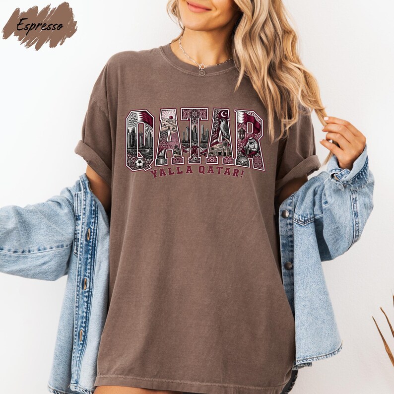 A woman wearing a brown t-shirt with the word "OAKAR" printed on it, along with the phrase "YALLA QATAR!" printed below. She is also wearing a denim jacket and has her arms outstretched.