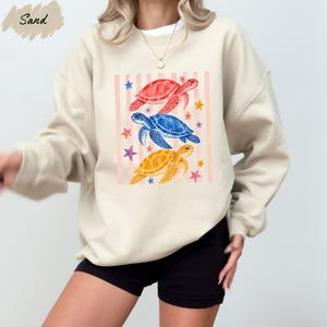 A woman wearing a beige sweatshirt with a colorful graphic design of three sea turtles and stars on it.