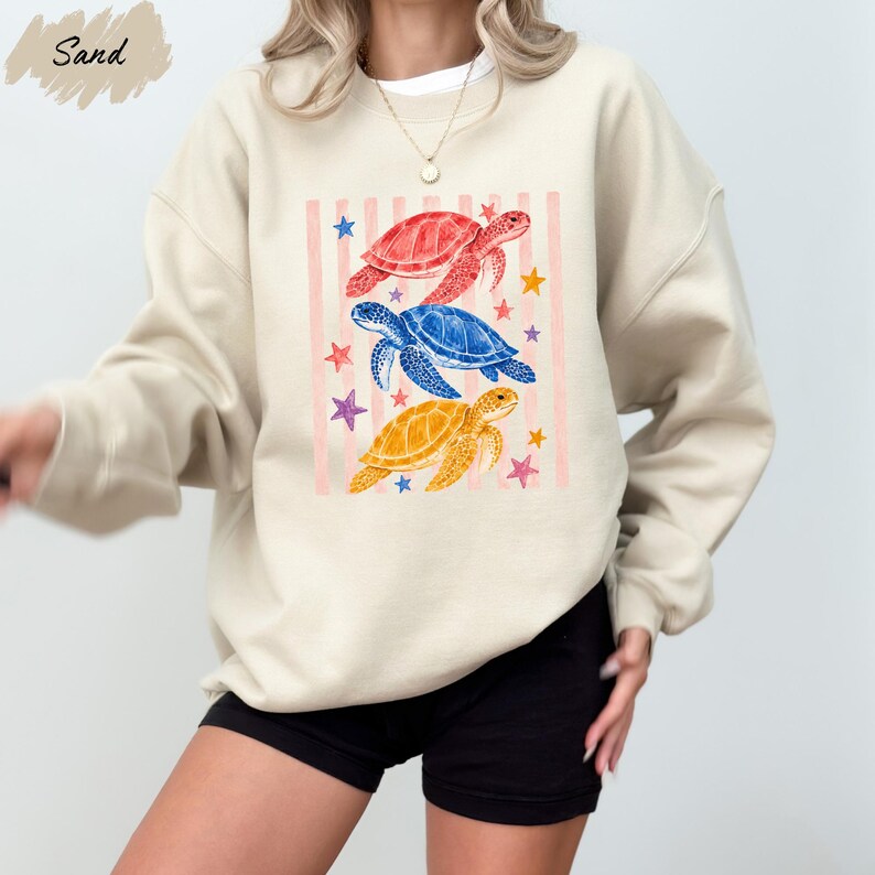 A woman wearing a beige sweatshirt with a colorful graphic design of three sea turtles and stars on it.