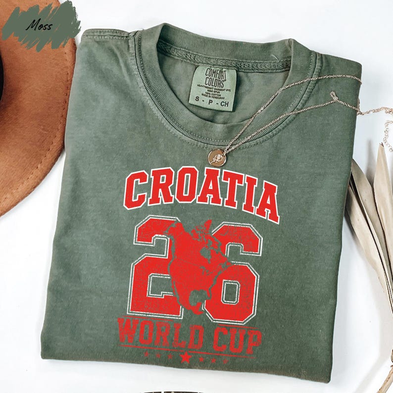 A green t-shirt with the text "Croatia 26 WORLD CUP" printed on it, along with a number "26" and a red graphic. The t-shirt is folded and placed on a white surface, with a brown hat and some leaves in the background.